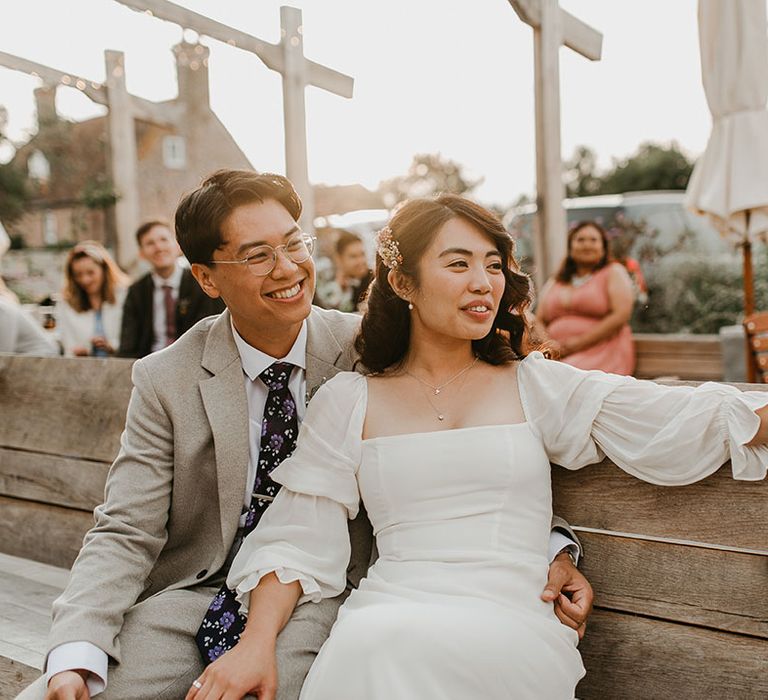 Bride & groom sit with one another on wooden bench outdoors on their wedding day