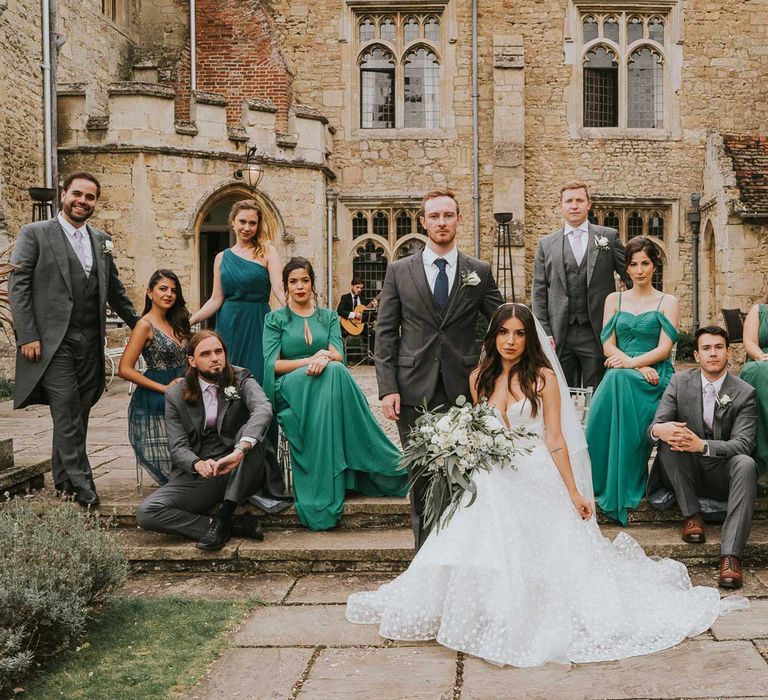 Bride in applique Pronovias wedding dress holding white and green bridal bouquet sits on step by standing groom in grey three piece suit surrounded by bridesmaids in assorted green bridesmaid dresses and groomsmen in grey morning suits in the grounds of Notley Abbey