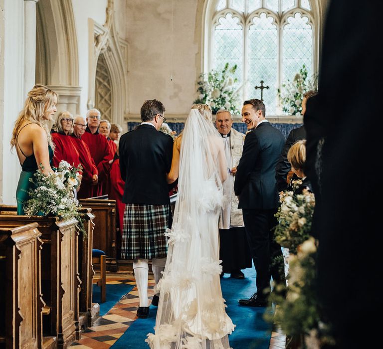 Bride in Halfpenny London wedding dress and ruffle Halfpenny London cathedral veil stands with groom in black tie at alter in church during wedding ceremony