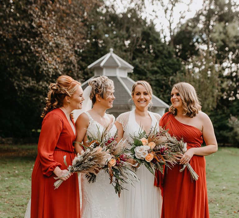 Brides stand with their bridesmaids who wear deep red gowns with Autumnal vibes and hold bright floral bouquets complete with pampas grass