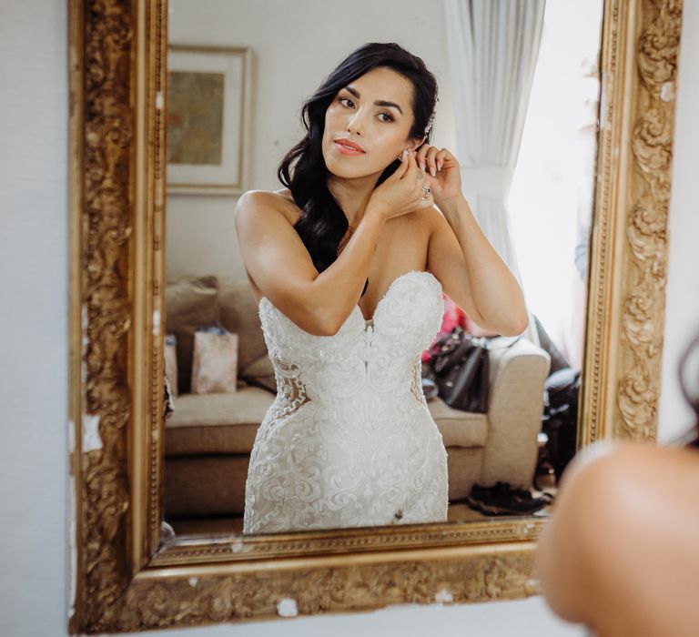 Bride in lace and tulle wedding dress with sweetheart neckline looks in the mirror whilst putting in her earrings 