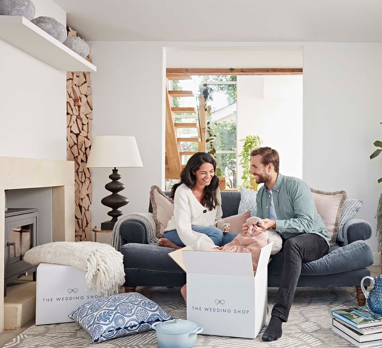 Couple sitting in their lounge on a blue plush velvet sofa opening their The Wedding Shop wedding gifts