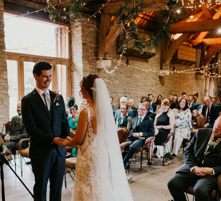 Bride in open back lace wedding dress and veil stands with groom in dark suit and blue tie during rustic wedding ceremony