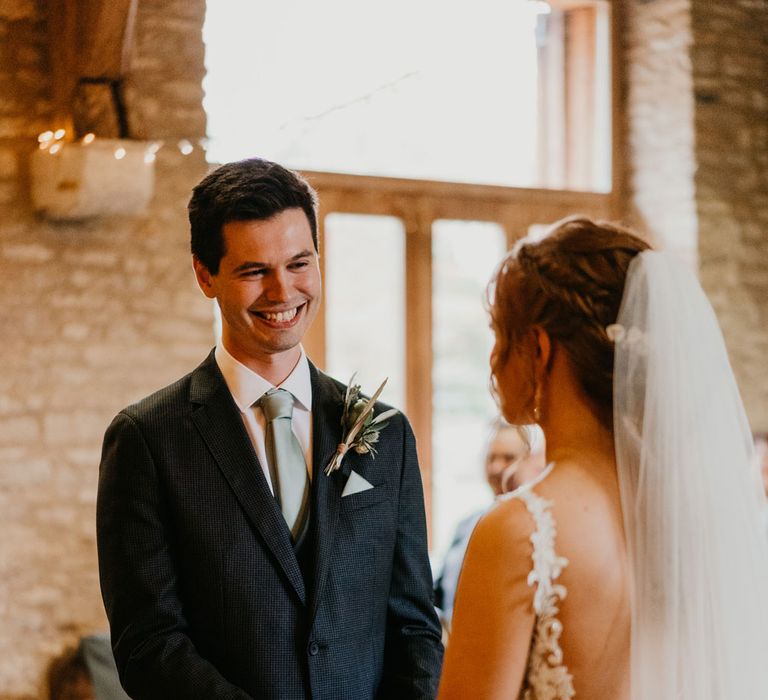 Smiling groom in dark suit, light blue tie and pocket square stands holding hands with bride in lace wedding dress during wedding ceremony