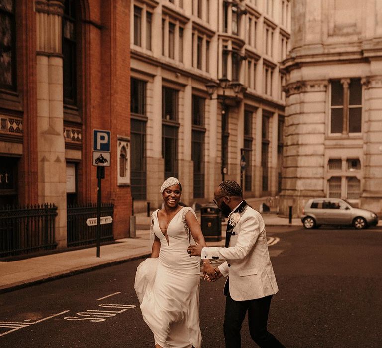 Groom in a white tuxedo jacket dancing with his bride in a fitted wedding dress and embellished cap headdress in the streets of Birmingham 