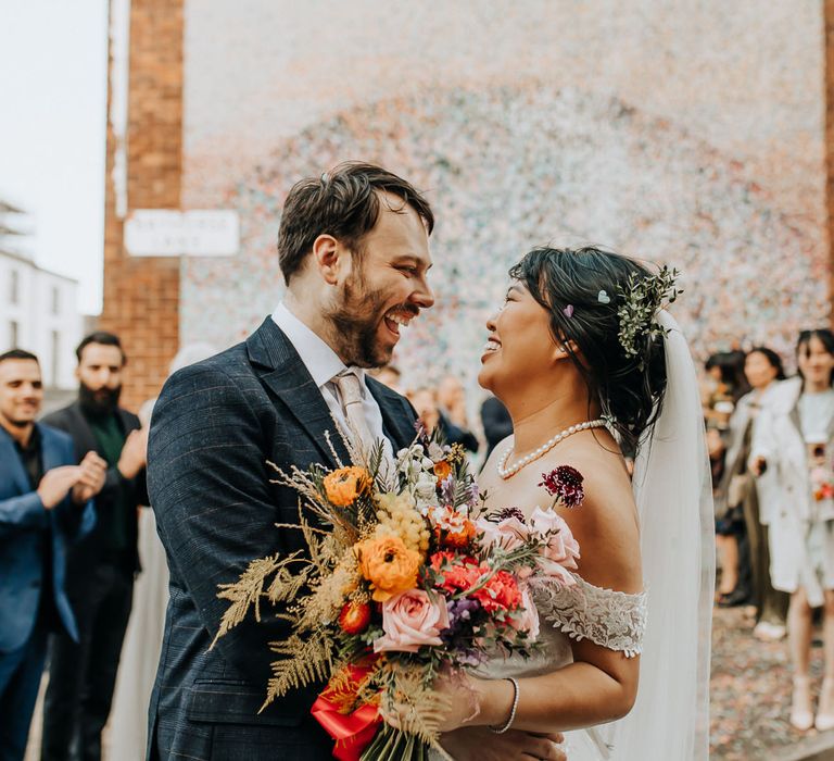 Smiling bride in pearl necklace and veil holding wild mixed wedding bouquet stands whipping groom in blue check suit with light pink tie after confetti toss