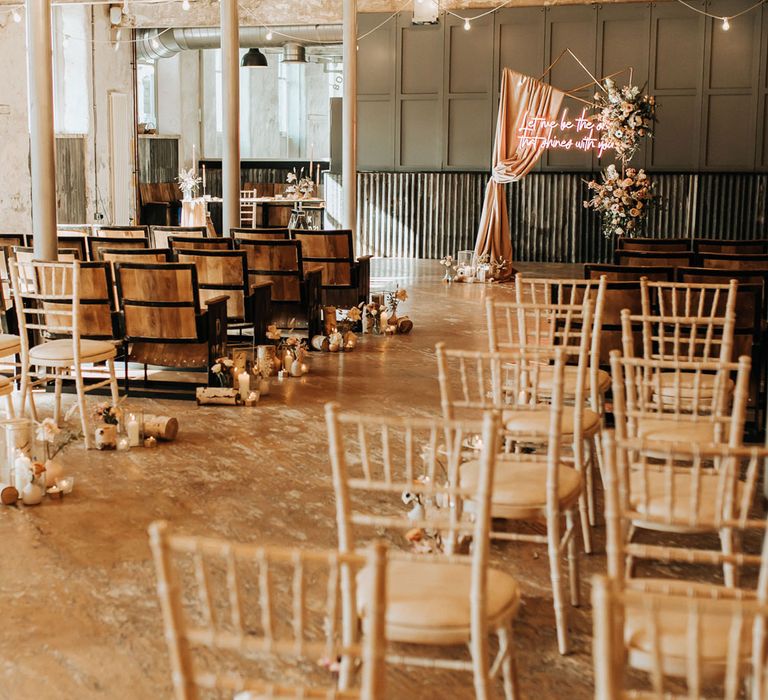 Interior of industrial boho wedding ceremony with pastel florals, bulb string lights and wooden chairs