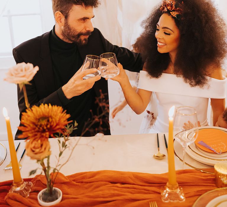 Black bride in an off the shoulder wedding dress with afro hair and hair accessory clinking glasses with her groom in a black suit 