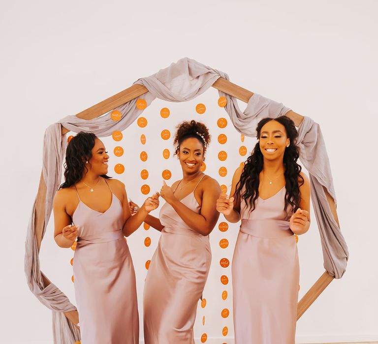 Bridal party dancing in rose satin bridesmaid dresses in front of a wooden frame escort card display 