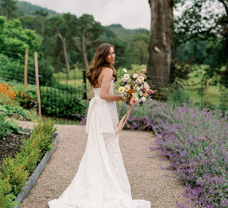 Bride with long brown curled hair holding wildflower bouquet in strapless lace wedding dress with bow