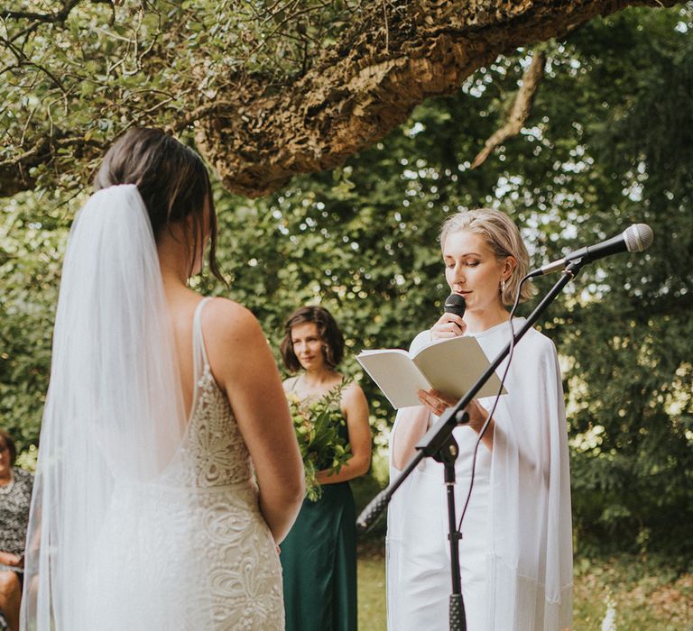 Bride in handmade jumpsuit with cape reads out wedding vows at outdoor wedding ceremony