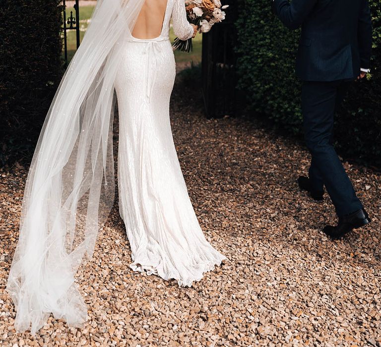 Bride in beaded dress with low back and bow with pearl veil walks along with groom in dark blue suit 