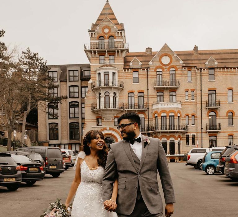 Bride in white traditional wedding dress and mixed flower neutral bouquet with groom in grey suit and black bow tie walking at The Petersham Hotel in Richmond