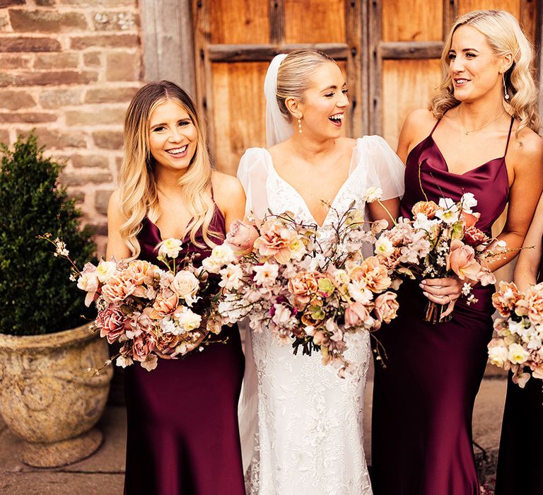 Bride in lace wedding dress stands with bridesmaid in burgundy satin cowl neck bridesmaid dresses 