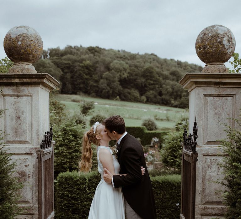 Groom in morning suit with grey trousers kisses bride in the gardens at Owlpen Manor 