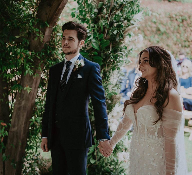 Bride in strapless sparkly wedding dress with detachable sleeves holds hands with groom in blue suit and polka dot tie during their wedding ceremony 