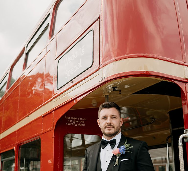 Groom stands on the double decker red bus wedding transport in black tie with a colourful wildflower buttonhole 