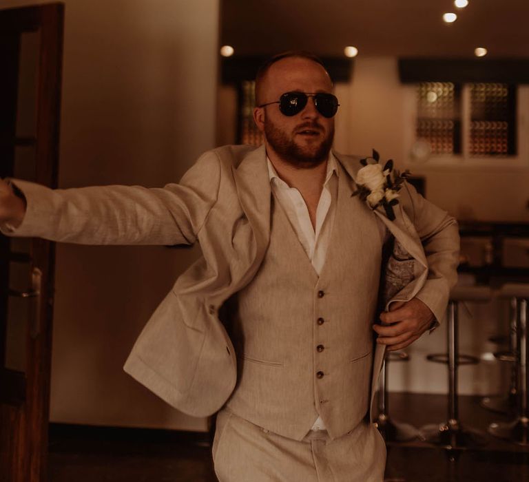 Groom wears three piece cream suit with open shirt and sunglasses and white floral buttonhole 