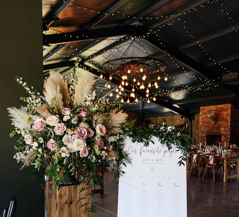 Traditional black and white wedding table plan sign with green foliage next to large pink rose and dried grass arrangement