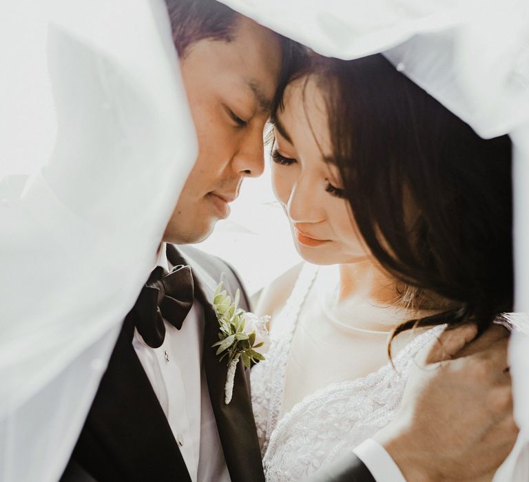 Close up image of bride & groom standing beneath pearl embellished veil 