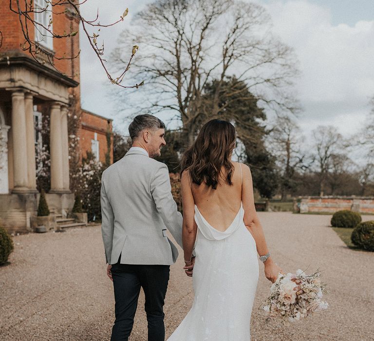 Groom in grey suit jacket walking with bride in backless satin wedding dress with custom embroidered train 