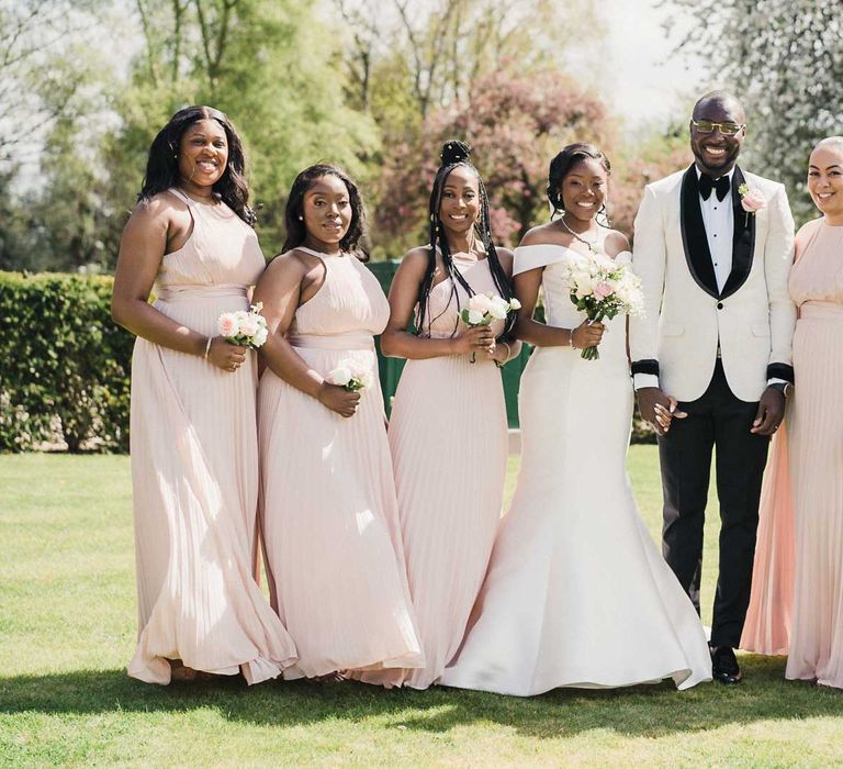 Bride & groom stand with bridesmaids who wear pale pink halter-neck dresses with silk ties to the waist