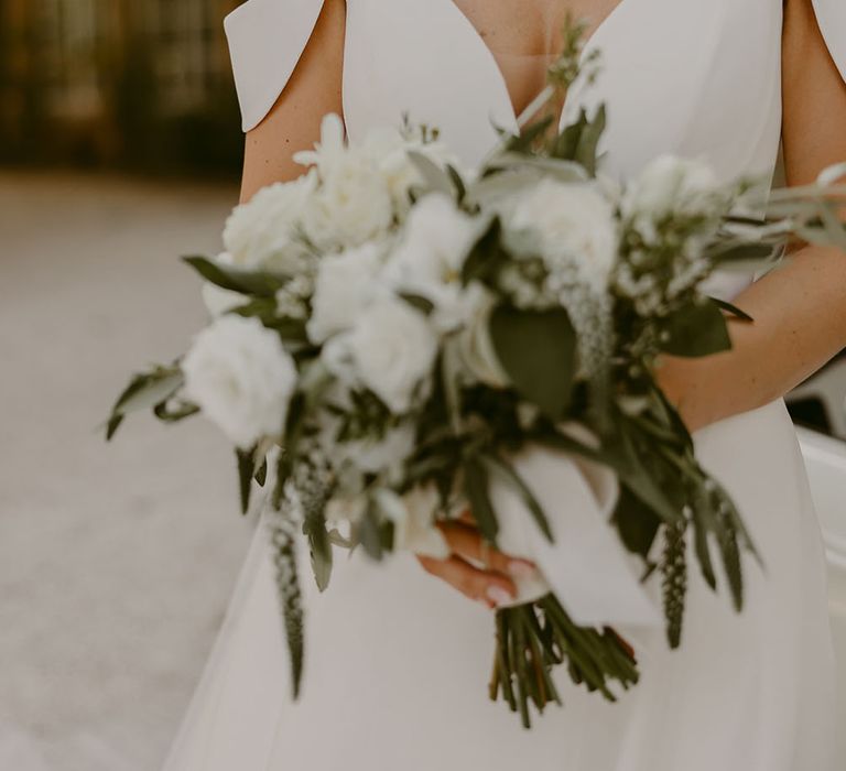 Bride wears plunge neckline Suzanne Neville wedding dress whilst holding white rose floral bouquet 