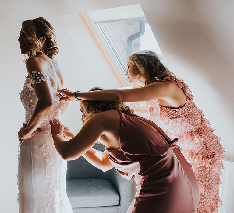 Bride in a leaf floral design off the shoulder wedding dress with the bridesmaids in pink dresses helping her out 