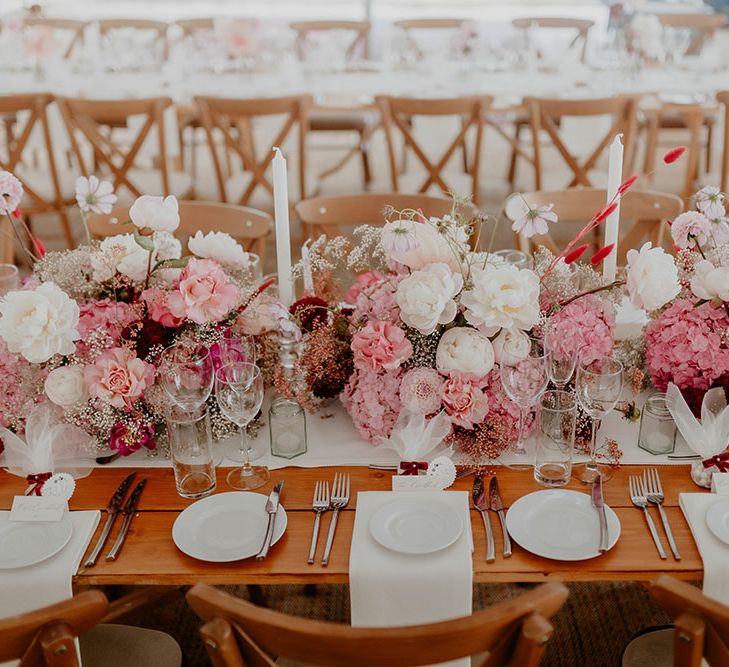 Wooden banquet tables with white fabric table runner with white and pink dalia flowers and noghl wedding favours 