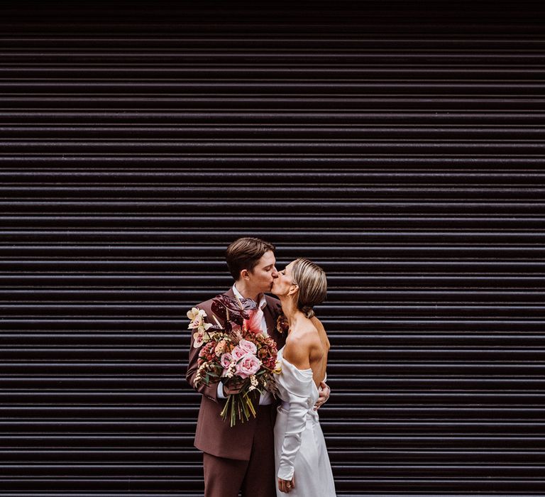 Groom holds the bridal bouquet as they share a kiss on their wedding day in London