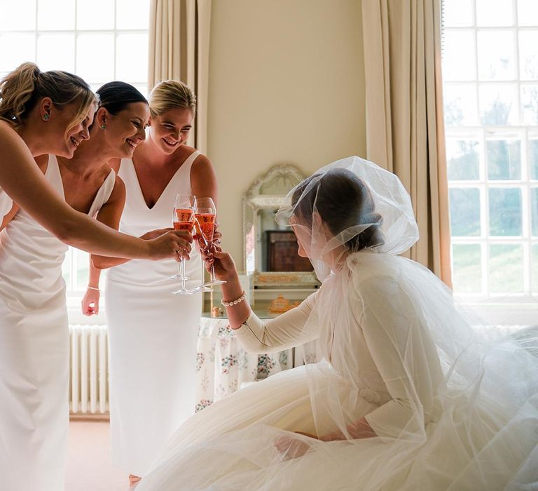 Bridesmaids wear white bridesmaid dresses and hold champagne glasses to cheers bride on the morning of her wedding day