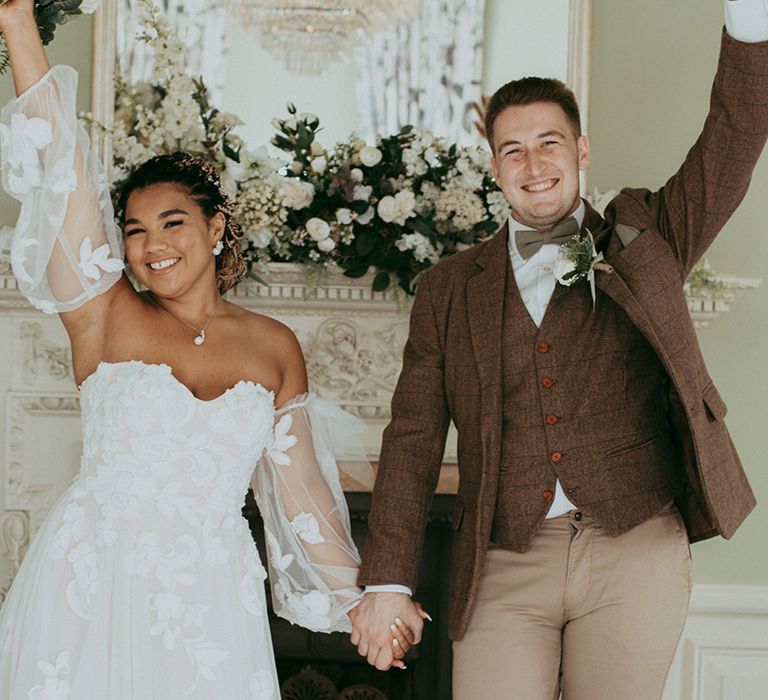 Bride in a strapless sweetheart wedding dress with sheer puff sleeves celebrates with the groom in a brown checkered suit 