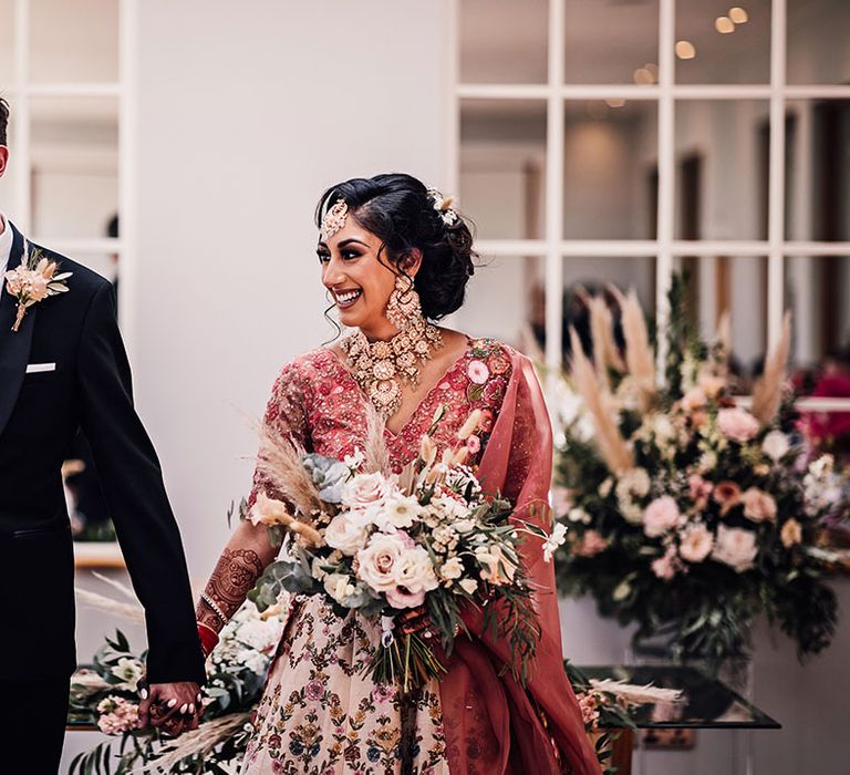 Bride carries neutral floral bouquet as she walks with her groom who wears black tie and pampas grass buttonhole 