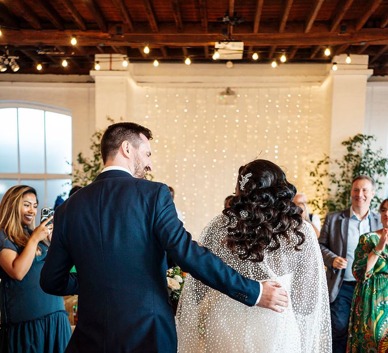 Bride wears embellished hair accessory in her black hair as she walks towards fairy light canopy at dry-hire venue