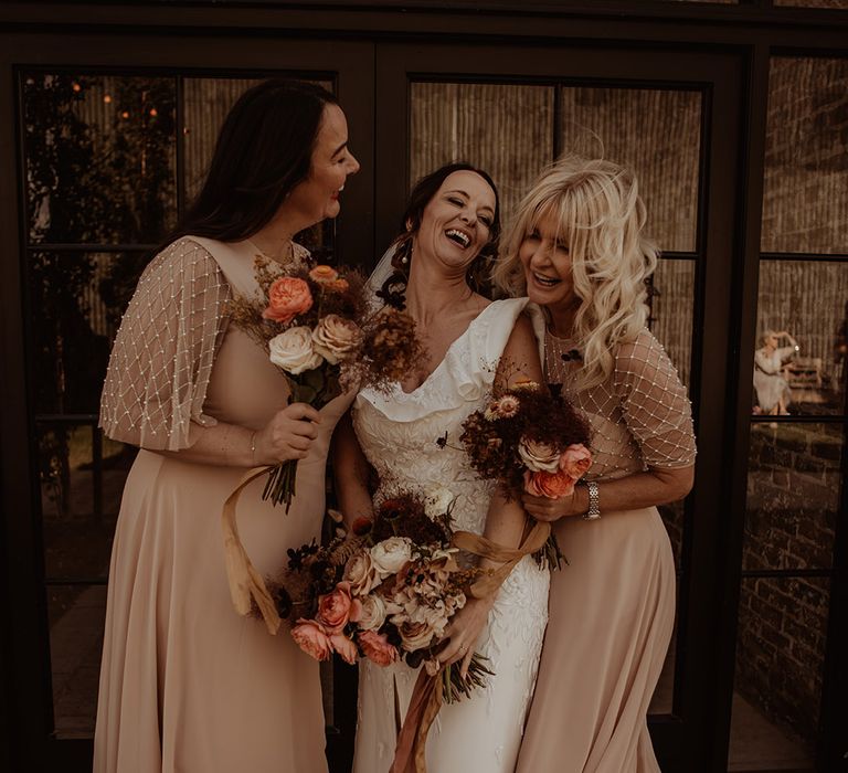 The bride laughs with her bridesmaids who wear blush pink dresses with pearl and beaded details 