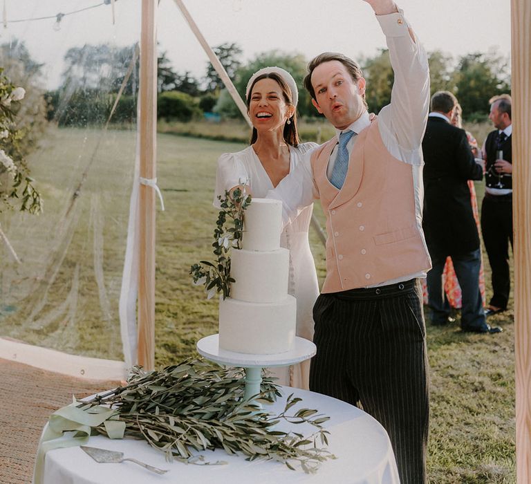 Three tier traditional white iced wedding cake with the bride in a satin button wedding dress and groom in pink waistcoat 