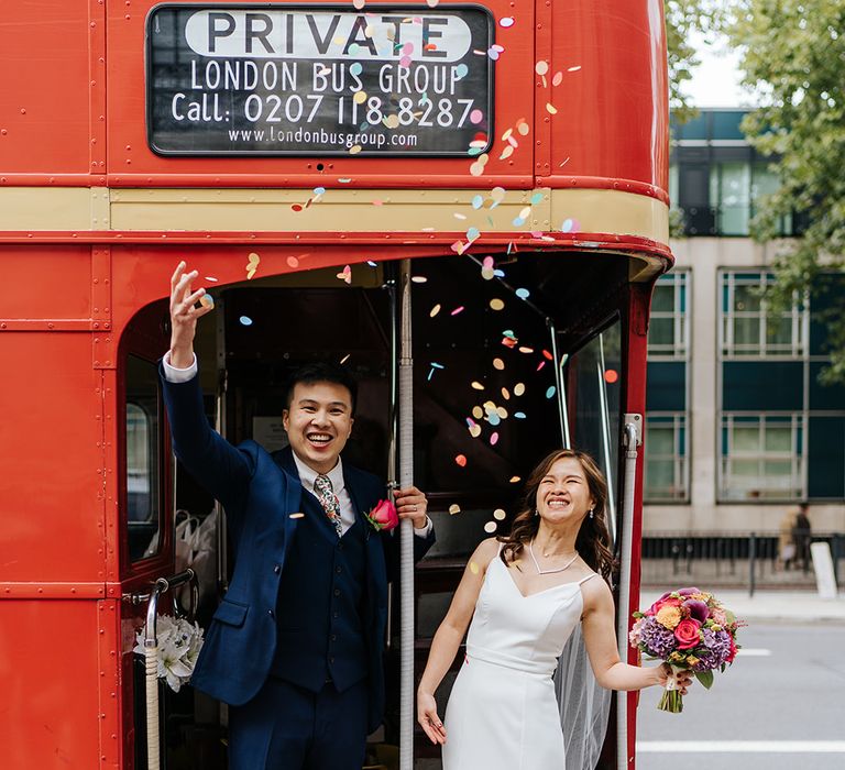 Groom throws colourful confetti into the air beside his bride on red London bus 