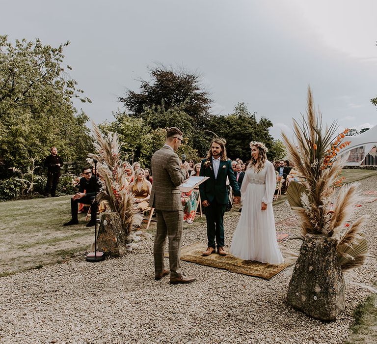 Bride & groom stand between dried floral and pampas installations outdoors during ceremony 