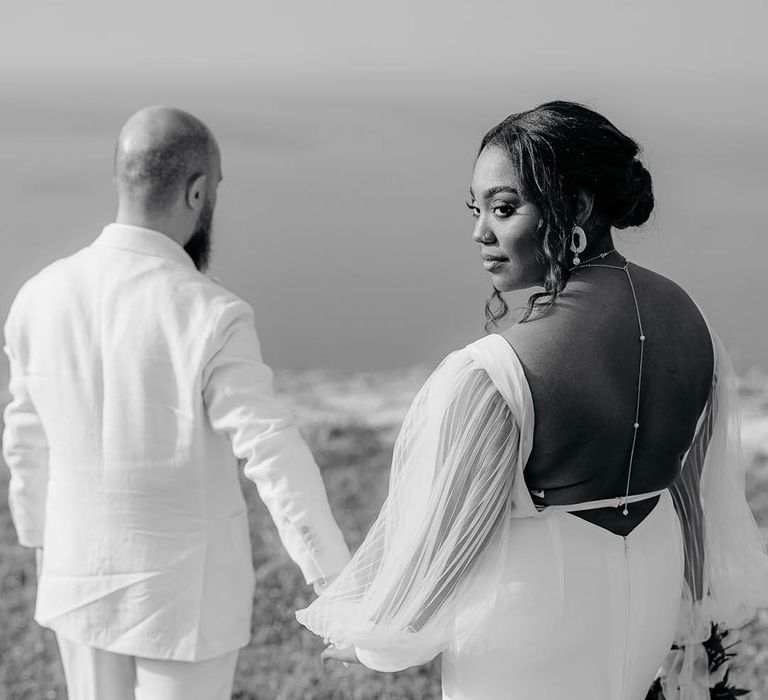 Bride looks back whilst holding the Grooms hand. She shows off her backless wedding dress and pearl jewellery