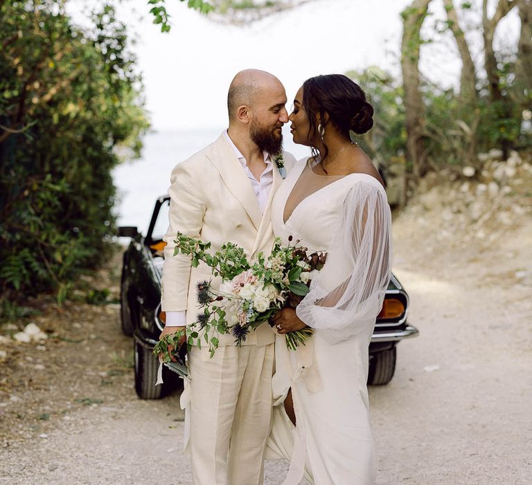 Bride and Groom look lovingly at each other in front of vintage Alfa Romeo Spidee. They wear a cream suit and sheer-sleeved wedding dress with Bride’s something blue wedding shoes on show