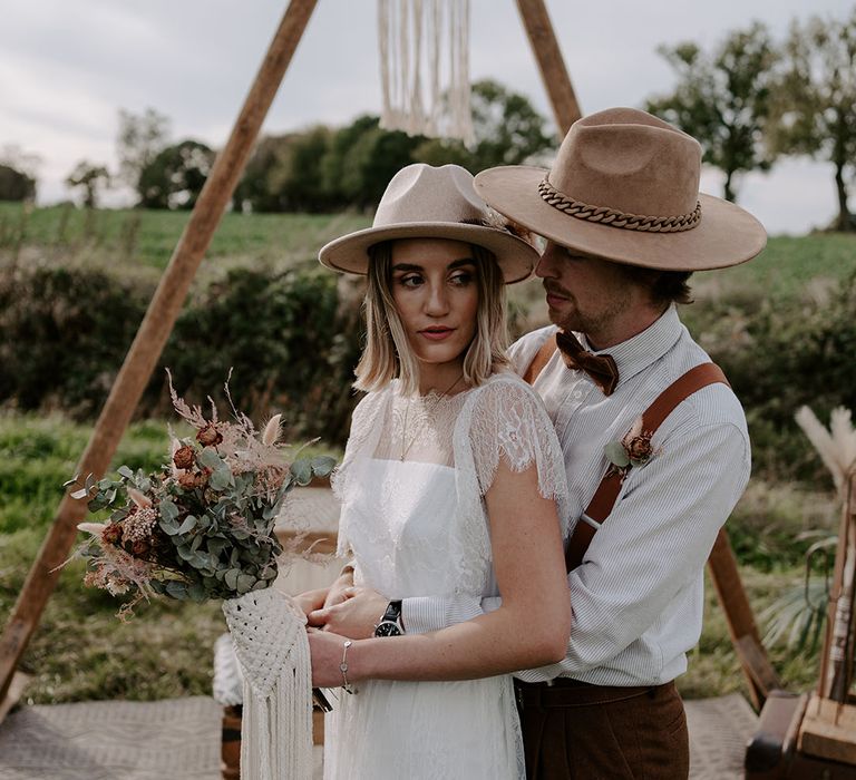 Groom in a striped shirt and brown hat with a chain embracing the bride in a boho wedding dress and beige hat with dried flowers 