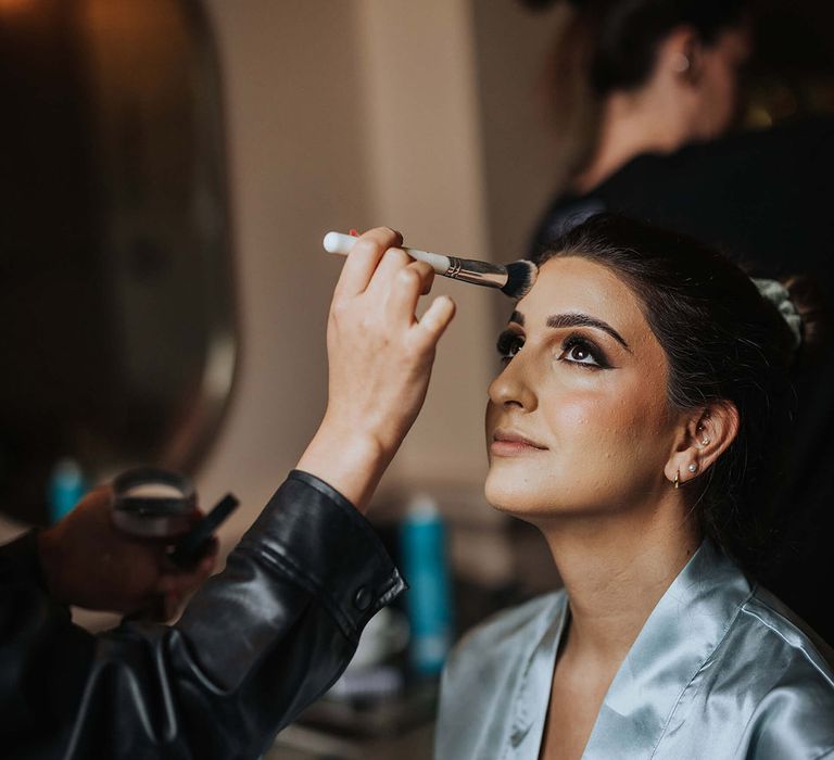 Bride in blue satin robe sits to get her makeup done with a smoky eye look