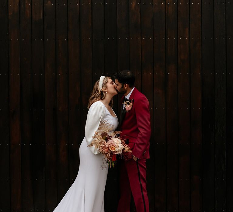 Bride in a pearl headband and button back wedding dress kissing the groom in a burgundy suit 