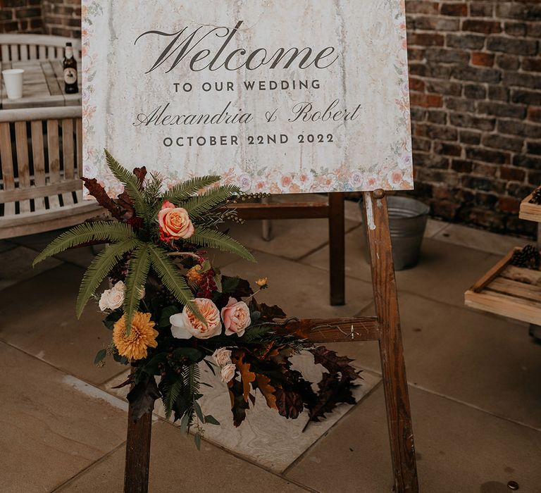 Rustic wedding welcome sign with flower designs around the edges on a wooden easel and autumnal flowers 