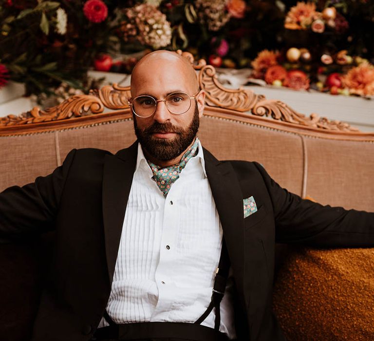 Groom on vintage orange loveseat wearing classic black tuxedo and black suspenders with hand-painted silk cravat and handkerchief 