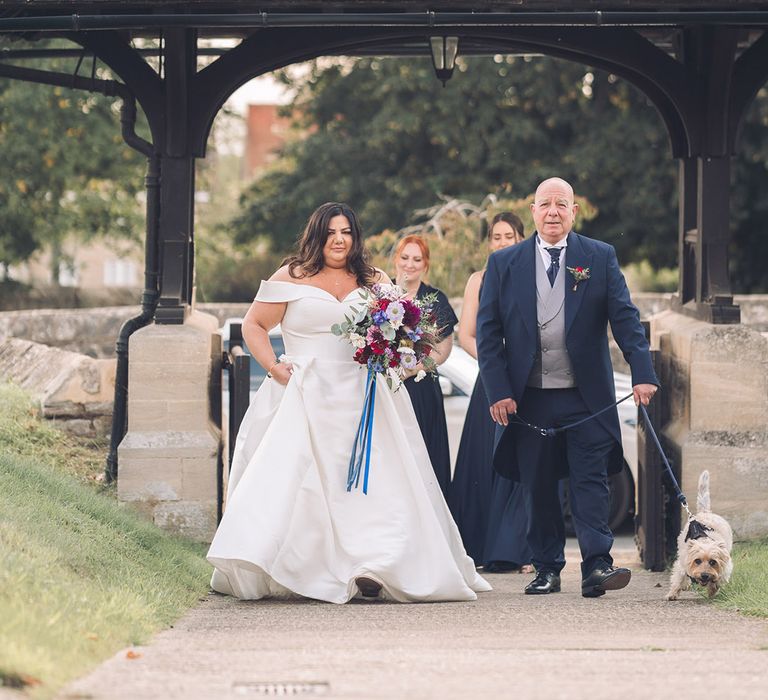 Bride in an off the shoulder wedding dress with purple and blue wedding bouquet walking with the father of the bride in a navy morning suit with the pet dog in a bow tie 