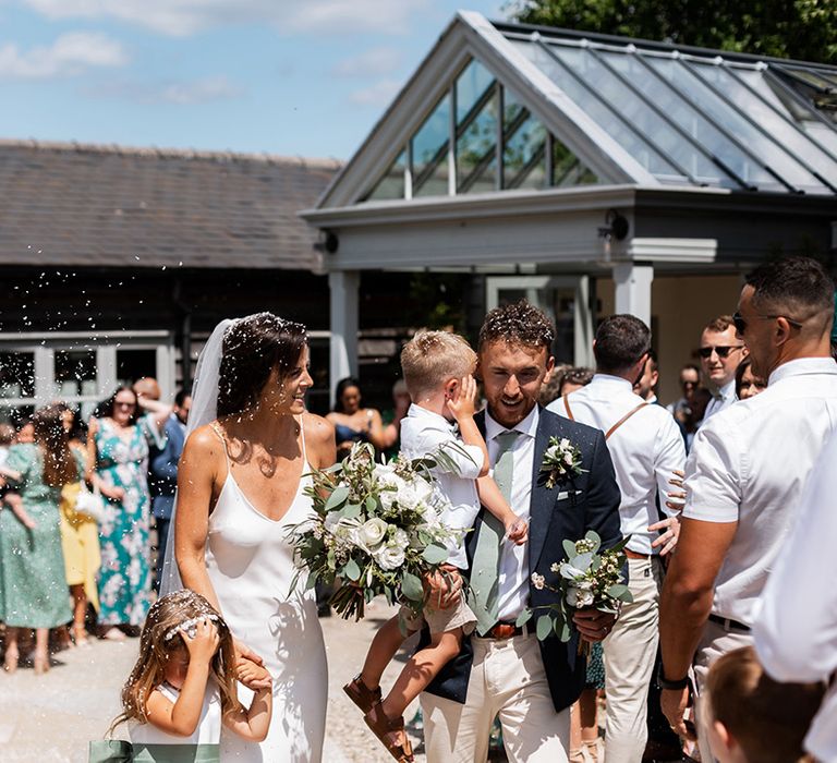 Bride in silk Grace Loves Lace wedding dress leaves wedding ceremony with her groom and children as white confetti is throw around them