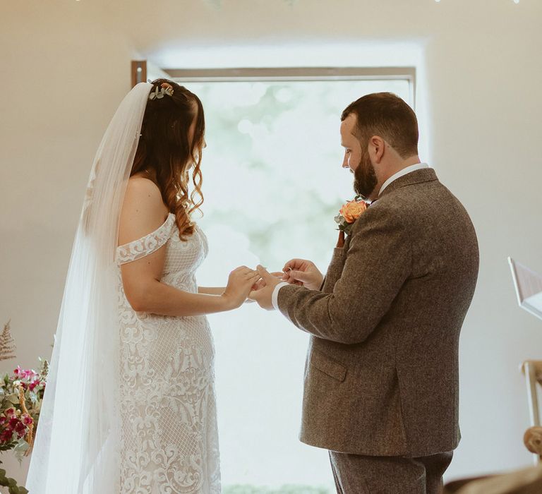 Groom in three piece tweed suit and orange buttonhole puts the ring on the bride's finger wearing a boho fitted off the shoulder lace wedding dress