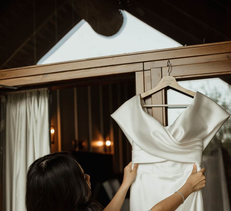 Bride with black hair in loose Hollywood waves in a white satin robe checking her off the shoulder Eve Lendel wedding dress 