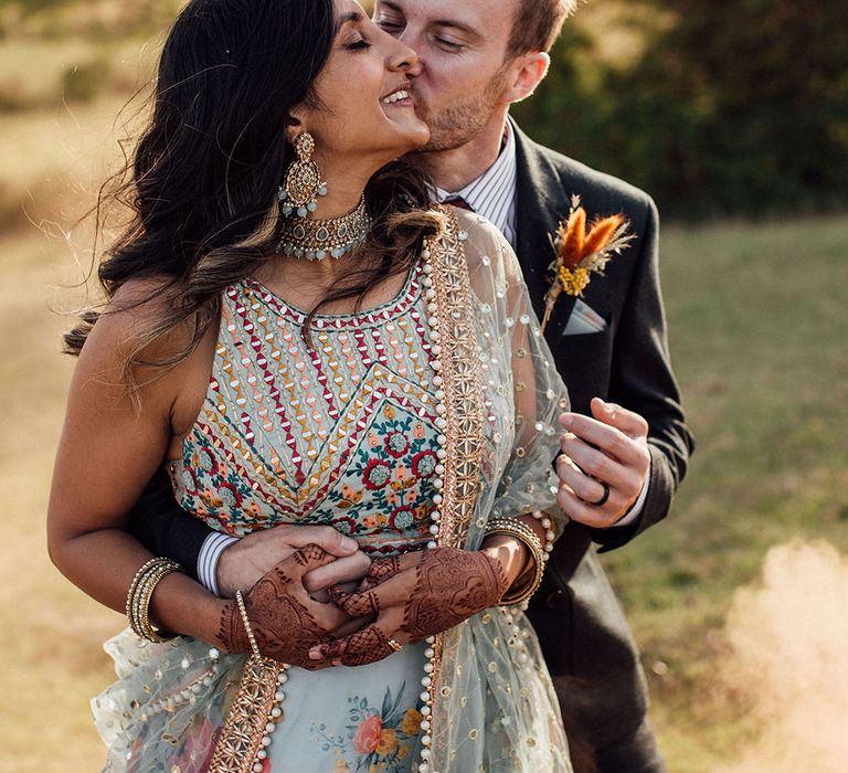 Groom wearing suit and colourful floral buttonhole embraces his bride in lehenga 